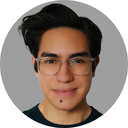 A close-up photo of a young man
										with a warm complexion, sporting sleek,
										dark hair styled upward. They are
										wearing clear-framed glasses and a
										dark, casual top. The person has a
										subtle smile and a beauty mark just
										below the lower lip. The background is
										simple grey color.