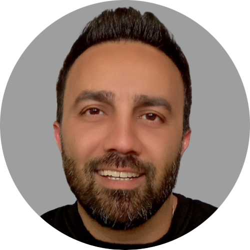 A close-up portrait of a man with
										an olive complexion, dark eyes, and a
										full beard. They have short, dark hair
										that is styled back and are smiling
										with a closed mouth. The individual is
										wearing a black top and the background
										is a simple grey color.